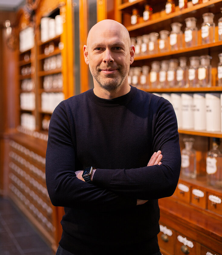 Mann mit Glatze und verschräkten Armen vor Apothekenregal.